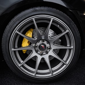 May include: A close-up shot of a car wheel featuring a silver alloy rim and a bright yellow brake caliper. The tyre is black, and the wheel has a multi-spoke design. The car's body is black.