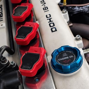 Puede incluir: Primer plano de un motor de coche con bobinas de encendido rojas y un tapón de aceite de motor azul. Los componentes del motor están dispuestos sobre una superficie metálica plateada. El texto "ENGINE OIL" es visible en el tapón.