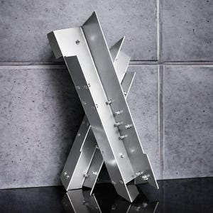 Könnte beinhalten: Eine moderne Metallskulptur im industriellen Stil. Die Skulptur besteht aus sich kreuzenden, abgewinkelten, silberfarbenen Metallträgern, die mit sichtbaren Schrauben befestigt sind. Das Design ist geometrisch und abstrakt, passend für moderne Dekoration.