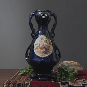May include: A dark blue ceramic vase with two handles and a decorative oval panel featuring a painted scene of figures. The vase is placed on a patterned rug with a small decorative box and greenery.