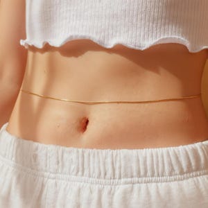 Könnte beinhalten: Eine zarte, goldfarbene Körperkette, die um die Taille getragen wird. Die Kette ist dünn und schlicht und verleiht einen subtilen Hauch von Eleganz. Das Model trägt ein weißes Crop-Top und eine weiße Jogginghose.