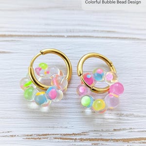 Könnte beinhalten: Handgefertigte goldene Creolen mit einem farbenfrohen Bubble-Bead-Design. Die Ohrringe haben goldene Reifen mit Clustern aus durchscheinenden, farbigen Perlen in Rosa, Grün, Gelb und Blau.