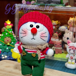 May include: A handmade crochet doll of Doraemon, wearing a red hat, striped shirt, green overalls, and a tan scarf. The doll has a white face with black eyes, a red nose, and pink whiskers. The text "Little Blue Chubby Doraemon" is at the bottom.