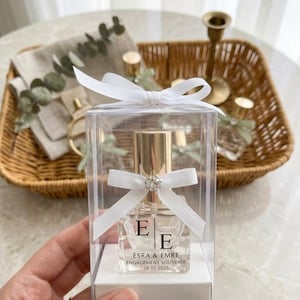 Può includere: Un flacone di profumo trasparente con tappo dorato è presentato in una scatola trasparente, legato con un nastro bianco e un piccolo gioiello. Il flacone ha le iniziali "E E" e il testo "ESRA & EMRE ENGAGEMENT SOUVENIR 28.03.2026".