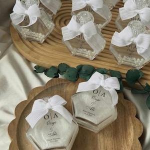 May include: Hexagonal glass bottles with white bows and pearl beads. Each bottle has text that reads "Özge & Ali 14.12.2026". The bottles are displayed on a wooden tray.