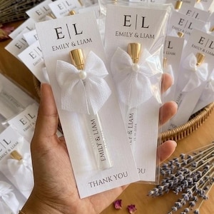 Pode incluir: Um frasco de perfume de vidro transparente com tampa dourada e um laço de tule branco com uma pérola central, fixado num cartão branco impresso com E|L, EMILY & LIAM e THANK YOU. O frasco tem impresso EMILY & LIAM e a data 12.10.2024.