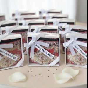May include: A collection of small gift boxes, each with a white lid and ribbon. Inside each box is a patterned textile. The textiles feature red, black, and beige designs. Each box has a tag that reads "Sarah's Henna Night".