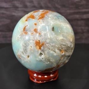 May include: A polished spherical stone featuring a light blue and translucent white base with scattered patches of rusty orange and brown mineral inclusions, resting on a small, circular, dark reddish-brown wooden display stand.