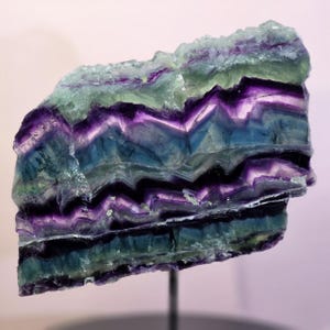 May include: A raw fluorite mineral specimen featuring distinct horizontal bands of deep purple, vibrant blue, and pale green. The stone displays a jagged, crystalline texture with sharp, angular layers and is mounted on a thin metal rod.
