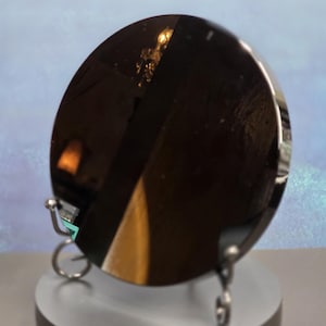 May include: A circular, highly reflective black mirror surface mounted on a silver-toned metal stand. The dark, polished disc features a smooth, glossy finish that creates a deep, dark appearance while reflecting light and surrounding objects.