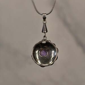 May include: A silver-toned pendant necklace featuring a circular, bowl-shaped setting with a small, round, purple gemstone centered at the base. The pendant hangs from a thin silver chain by a ribbed, conical bail connector.