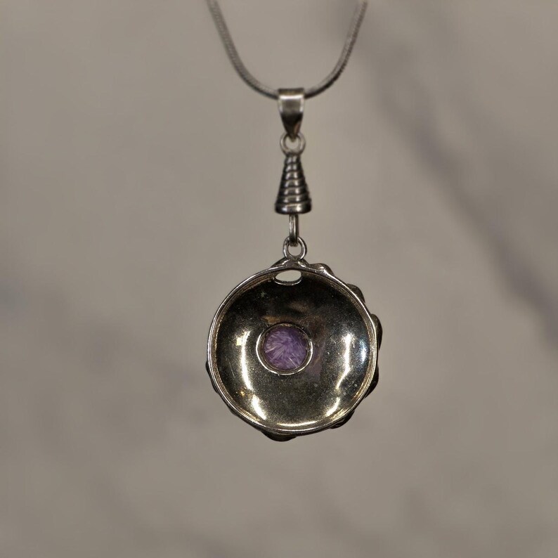 May include: A silver-toned pendant necklace featuring a circular, bowl-shaped setting with a small, round, purple gemstone centered at the base. The pendant hangs from a thin silver chain by a ribbed, conical bail connector.