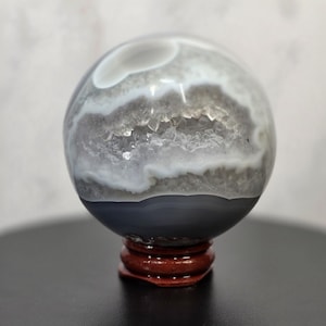 May include: A polished agate sphere featuring a central geode cavity filled with sparkling white quartz crystals. The stone displays concentric bands of grey, white, and translucent layers, resting on a small, circular, dark reddish-brown wooden stand.