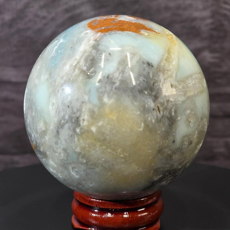 May include: A polished spherical stone featuring a marbled pattern of light blue, grey, white, and earthy orange tones. The smooth, rounded mineral orb rests securely on a small, circular, dark reddish-brown wooden display stand.