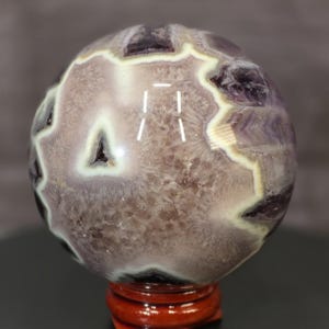 May include: A polished spherical amethyst crystal featuring deep purple zones, translucent white quartz bands, and a textured, crystalline interior surface. The sphere is displayed on a small, circular, reddish-brown wooden stand.