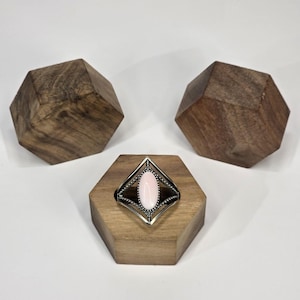 May include: A silver ring featuring a light pink oval gemstone within a diamond-shaped setting. The ring is presented on a wooden hexagonal stand, with two wooden geometric shapes in the background.