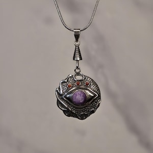 May include: A silver pendant necklace featuring a circular design with a central purple stone shaped like an eye. The metal surface has a textured, honeycomb-like pattern and includes three small orange circular accents above the eye motif.