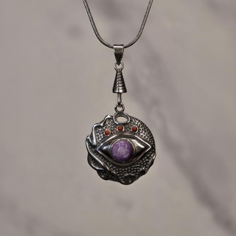 May include: A silver pendant necklace featuring a circular design with a central purple stone shaped like an eye. The metal surface has a textured, honeycomb-like pattern and includes three small orange circular accents above the eye motif.