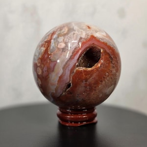 May include: A polished agate sphere featuring a mix of deep red, brown, and translucent white banding. The stone has a prominent, jagged opening on its side revealing a dark, crystalline interior. It rests on a small, circular red stand.