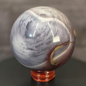 May include: A polished spherical stone featuring swirling patterns of grey, white, and deep purple. The surface displays a smooth, reflective texture with a distinct golden-brown mineral inclusion along the side, resting on a small wooden stand.