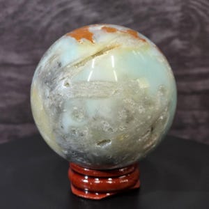 May include: A polished spherical stone featuring a mix of pale blue, translucent white, and earthy orange-brown patches. The surface displays natural crystalline inclusions, small crevices, and a smooth texture, resting on a small circular wooden stand.