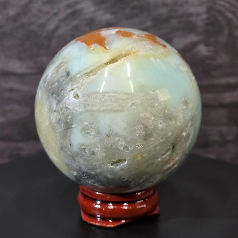 May include: A polished spherical stone featuring a mix of pale blue, translucent white, and earthy orange-brown patches. The surface displays natural crystalline inclusions, small crevices, and a smooth texture, resting on a small circular wooden stand.