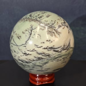 May include: A polished spherical stone featuring a pale mint green and off-white base color with intricate, dark brown dendritic fern-like inclusions spreading across the surface, resting on a small, circular, glossy dark red wooden stand.