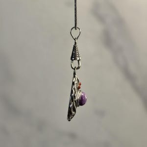 May include: A silver-toned pendant hanging from a thin chain, featuring a circular metal base with intricate wirework patterns. A smooth, round purple stone is set in the center, accented by small, textured red beads near the top bail.
