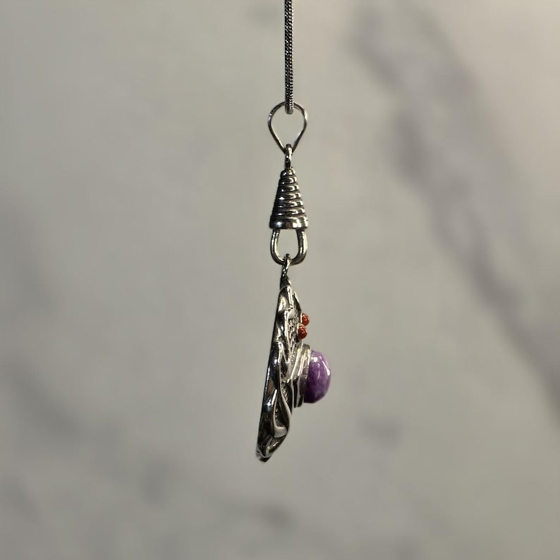 May include: A silver-toned pendant hanging from a thin chain, featuring a circular metal base with intricate wirework patterns. A smooth, round purple stone is set in the center, accented by small, textured red beads near the top bail.