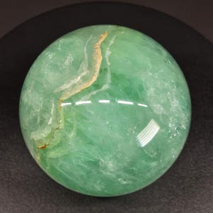 May include: A polished green fluorite sphere featuring translucent layers and internal crystalline structures. The surface displays a prominent, jagged vein of white and tan mineral inclusions that cut across the smooth, rounded gemstone.