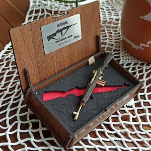 Könnte beinhalten: Eine Miniatur-Nachbildung eines M1-Karabiners aus dunklem Holz und Messing, in einer hölzernen Präsentationsbox mit schwarzer Schaumstoffeinlage und rotem Ausschnitt. Ein silbernes Metallschild im Deckel trägt den Text M1 CARBINE, semi-automatic carbine, 1941-Caliber 30.