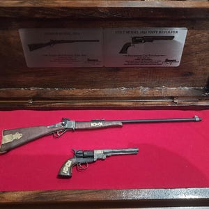 Könnte beinhalten: Eine hölzerne Präsentationsbox mit rotem Samtinnenfutter, die zwei Miniatur-Feuerwaffenmodelle enthält. Das obere Modell ist ein Sharps Model 1874 Gewehr, das untere ein Colt Model 1851 Navy Revolver. Metallschilder im Deckel zeigen Text und Abbildungen der jeweiligen Schusswaffen.