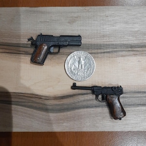 Puede incluir: Dos pistolas de juguete en miniatura con armazones negros y empuñaduras marrones colocadas junto a una moneda de cuarto de dólar estadounidense. La pistola superior es de estilo semiautomático y la inferior tiene un mecanismo de palanca.