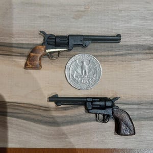 Könnte beinhalten: Zwei schwarze Miniatur-Revolver mit braunen Holzmaserungsgriffen, die über und unter einer US-Vierteldollarmünze platziert sind, welche als Größenvergleich für die geringe Größe der beiden Spielzeugschusswaffen dient.