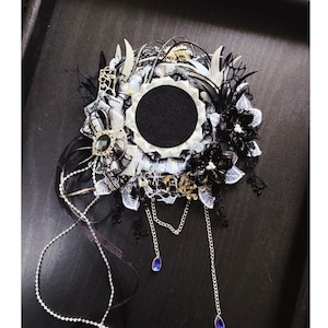 Könnte beinhalten: Ein gotischer Haarschmuck mit einem zentralen schwarzen Kreis, umgeben von weißer Spitze, schwarzen Federn und floralen Akzenten. Silberketten mit blauen Tropfenanhängern hängen von unten. Das Design weist aufwendige Details und eine dunkle Ästhetik auf.