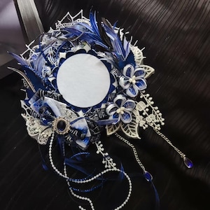 Puede incluir: Un tocado ornamentado con un círculo blanco central, rodeado de tela azul y blanca, encaje y plumas. El diseño incluye detalles florales, una broche enjoyada y cadenas colgantes con colgantes de lágrima azules. Un accesorio decorativo.