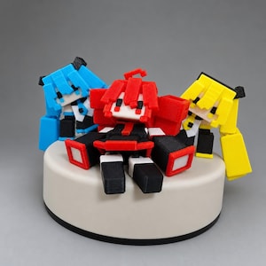 May include: Three blocky, pixel-style figurines featuring square heads and limbs, arranged on a circular white base. The central figure is red, the left figure is bright blue, and the right figure is yellow. Each character has black accents.