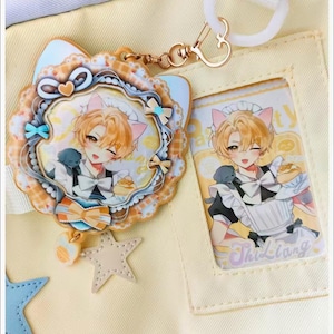 May include: A decorative keychain and card featuring an anime-style character with cat ears, holding food. The keychain has a layered design with a star charm. The card has the name "Thi Liang" printed on it. The background is a light yellow color.