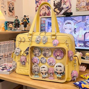 May include: A yellow satchel bag with a clear front pocket displaying character pins and figurines. The bag is adorned with character keychains. The bag is sitting on a wooden surface with manga books and figurines in the background.