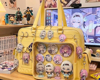 Puede incluir: Un bolso amarillo con un bolsillo frontal transparente que muestra pines y figuras de personajes. El bolso está adornado con llaveros de personajes. El bolso está sobre una superficie de madera con libros manga y figuras en el fondo.