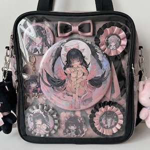 May include: Black tote bag with a clear front panel displaying anime-style character pins, charms, and rosettes in pink and black. A large circular pin features a winged character. A pink bow and a small pink teddy bear are attached.