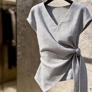 May include: A light gray short-sleeved wrap top featuring a V-neckline and a side tie closure. The garment is constructed with a textured fabric and hangs from a black hanger, showcasing a draped silhouette with an asymmetrical hemline.