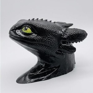 May include: A black dragon head figurine with a textured, scaly surface and a single prominent, bright lime green eye. The creature features elongated ear-like appendages extending from the back of the head and a series of small ridges along the top.