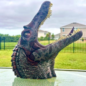 May include: A detailed dinosaur bust featuring a textured, dark reddish-brown and grey scaly skin pattern. The creature has an elongated snout with an open mouth, revealing rows of sharp, pale yellow teeth and a dark interior throat.