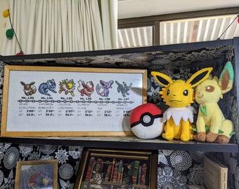 May include: A framed cross-stitch of the first generation Pokemon, Eevee and its evolutions, with a Pokeball and two plush Eevee toys. The cross-stitch includes the Pokemon's names, numbers, and stats. The plush toys are yellow and green.