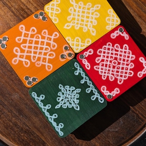May include: Four square coasters in bright colours: orange, yellow, red, and green. Each coaster features a white, intricate geometric design. Small silver accents are visible on each corner. The coasters are arranged on a wooden tray.