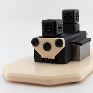 May include: A black and beige industrial-style object with a rectangular base. The object has two stacked, square, black components with floral-like cutouts. A beige, triangular piece with three black screws is on the front.