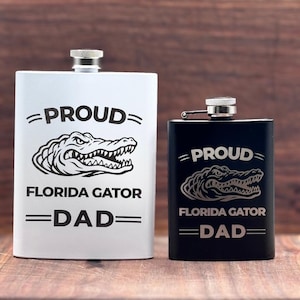 Może przedstawiać: Biała metalowa piersiówka i mniejsza czarna metalowa piersiówka, obie z grafiką głowy aligatora i napisem PROUD FLORIDA GATOR DAD. Każda piersiówka posiada srebrną zakrętkę oraz prostokątny korpus z zaokrąglonymi krawędziami.