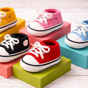 Peut inclure: Six chaussons de bébé au crochet faits à la main, de couleurs jaune, rose, bleu, noir et rouge. Chaque chaussure présente des lacets blancs, un bout blanc et une bande noire le long de la semelle. Les chaussures sont exposées sur des blocs géométriques colorés.