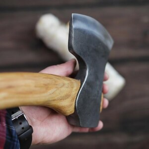 Puede incluir: Primer plano de una cabeza de hacha de acero con un mango de madera. La cabeza del hacha es de color gris oscuro y el mango de color marrón claro. El hacha se sostiene en una mano, con una camisa a cuadros y un reloj negro visibles en el fondo.